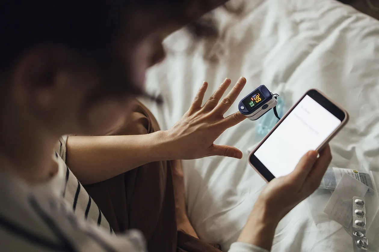 Woman sits on bed taking vital sign reading and looks at phone. 